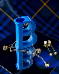 Burberry letter B blue candle draped with gold jewelry and earrings on a reflective surface against blue tartan.