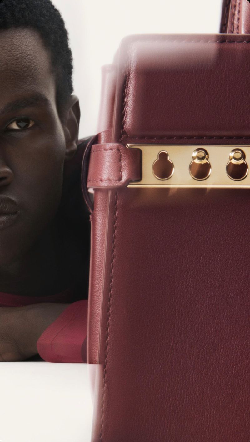 Close-up on a maroon leather handbag with gold hardware, partially obscuring a dark-skinned man wearing a maroon shirt, on a white surface.