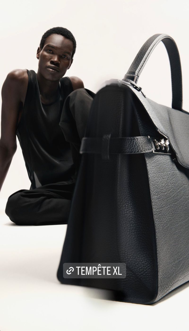 Dark-skinned man poses next to a large, structured black leather satchel handbag with white stitching, against an off-white backdrop.