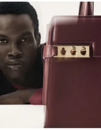 Close-up portrait of a Black male model peering past a large burgundy leather handbag with gold hardware