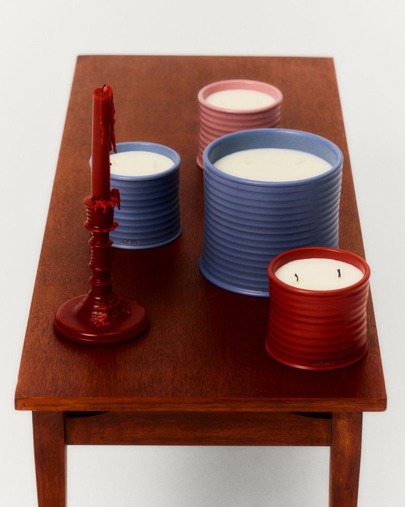 Still life shot of candles in colorful, ridged tins and a red candlestick on a brown wooden table against a white backdrop.