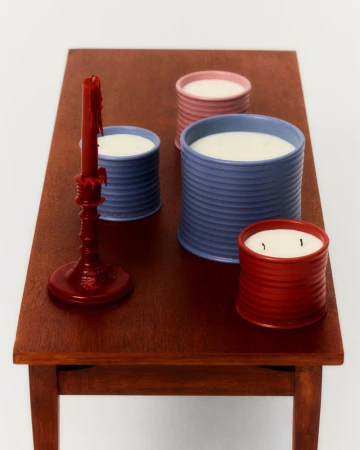 Still life shot of candles in colorful, ridged tins and a red candlestick on a brown wooden table against a white backdrop.