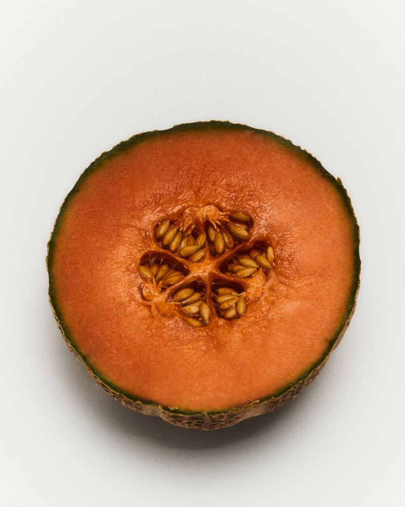 Studio shot of a halved cantaloupe melon on a white background showing the orange flesh and seeds in the center, simple food still life.