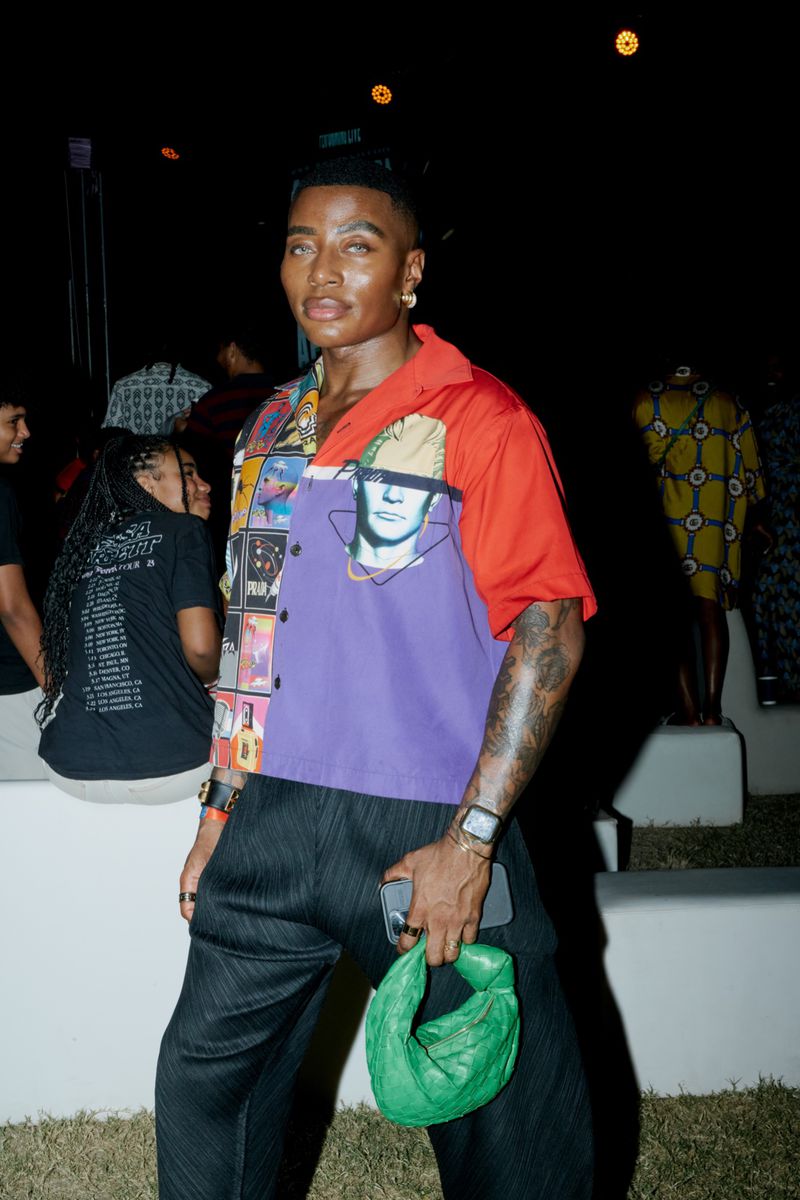 Stylish individual poses in colorful patterned shirt and black pants, holding green purse at nighttime event with blurred people in background.
