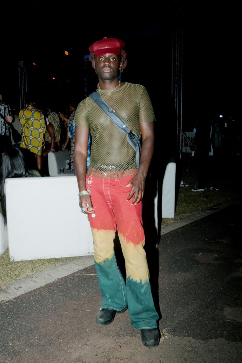 Person with mesh top and red, yellow and green pants posing outdoors at night, wearing a red cap and black shoes, with a satchel bag.