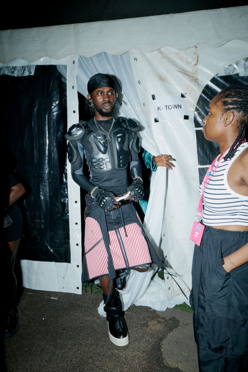 An individual in distinctive black armor and a skirt with red stripes stands outside a tent with another person in a striped shirt.