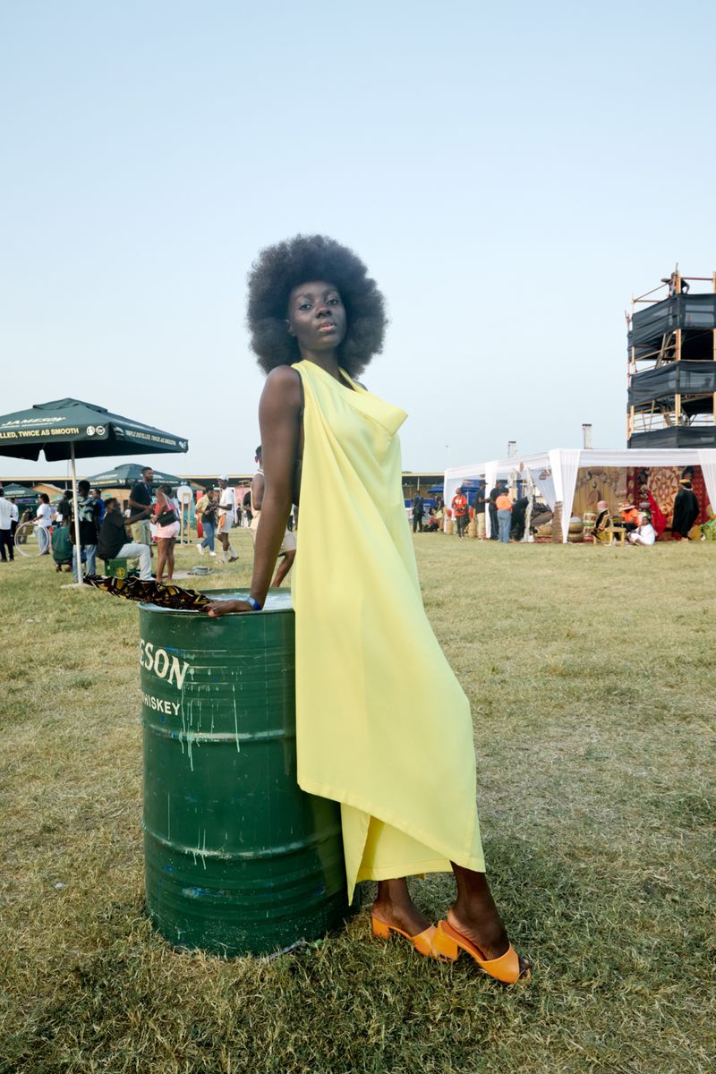 An adult female in a yellow dress poses next to a green metal barrel at an outdoor event with people and tents in the background.