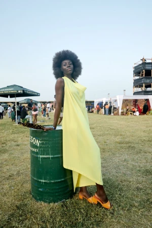 An adult female in a yellow dress poses next to a green metal barrel at an outdoor event with people and tents in the background.