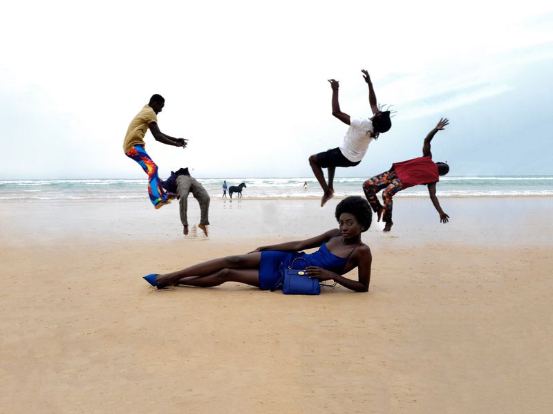 A dark-skinned woman in a blue dress poses on a beach with three men jumping in the background near the ocean water and a horse.