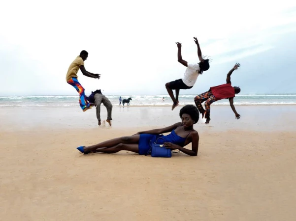 A dark-skinned woman in a blue dress poses on a beach with three men jumping in the background near the ocean water and a horse.