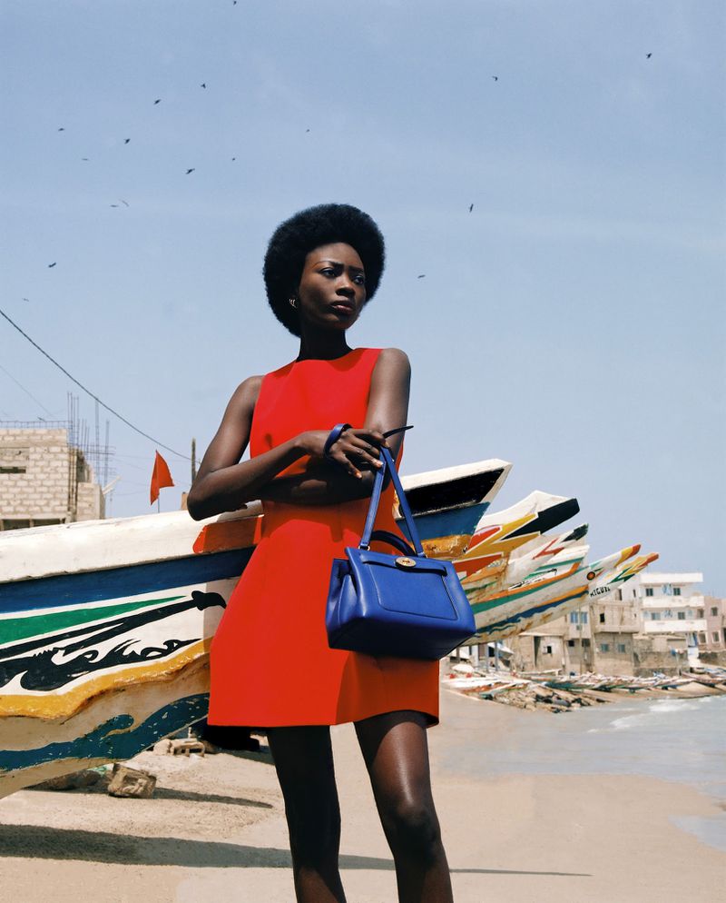 Portrait of a dark-skinned woman in a red dress, standing next to colorful fishing boats, holding a small blue handbag in an outdoor scene.