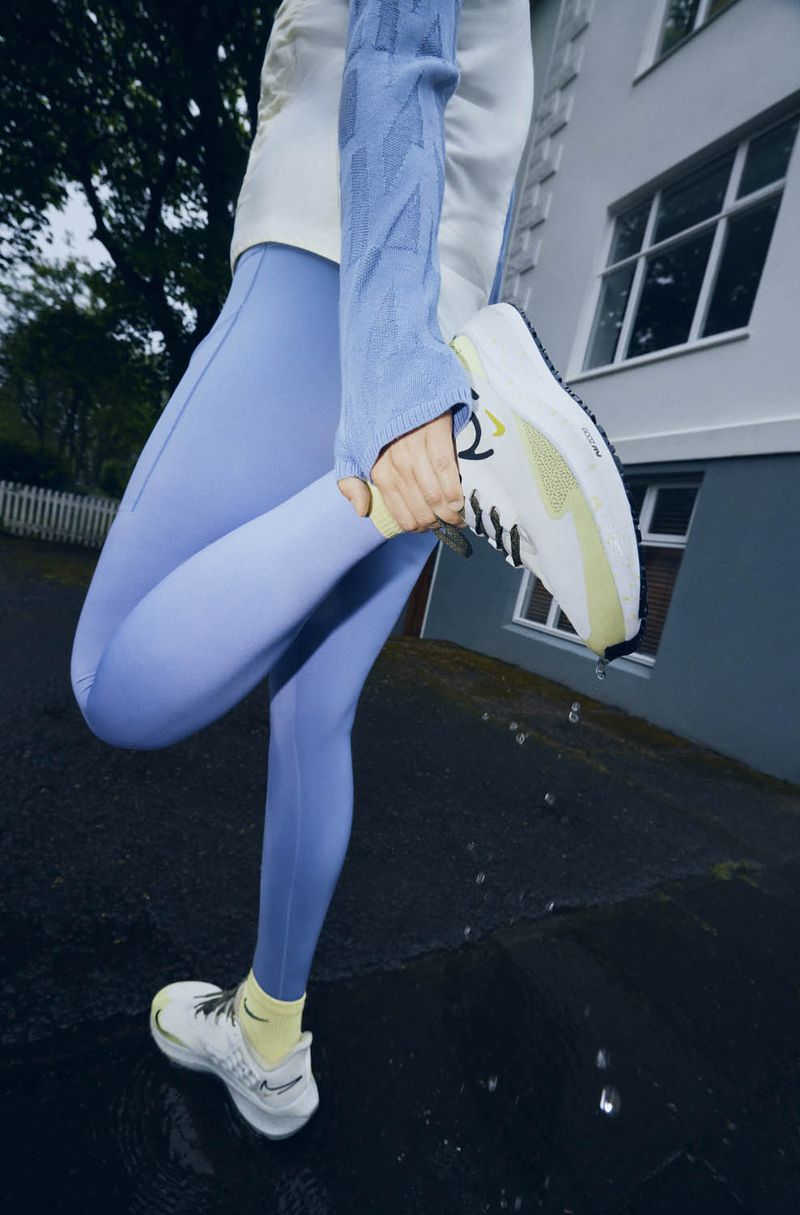 Athletic woman in blue leggings stretches her leg outdoors, showcasing white running shoe with black laces and a yellow accent.