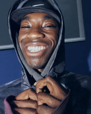 Close-up portrait of a smiling Black man wearing a dark hooded jacket and cap against a plain blue background.