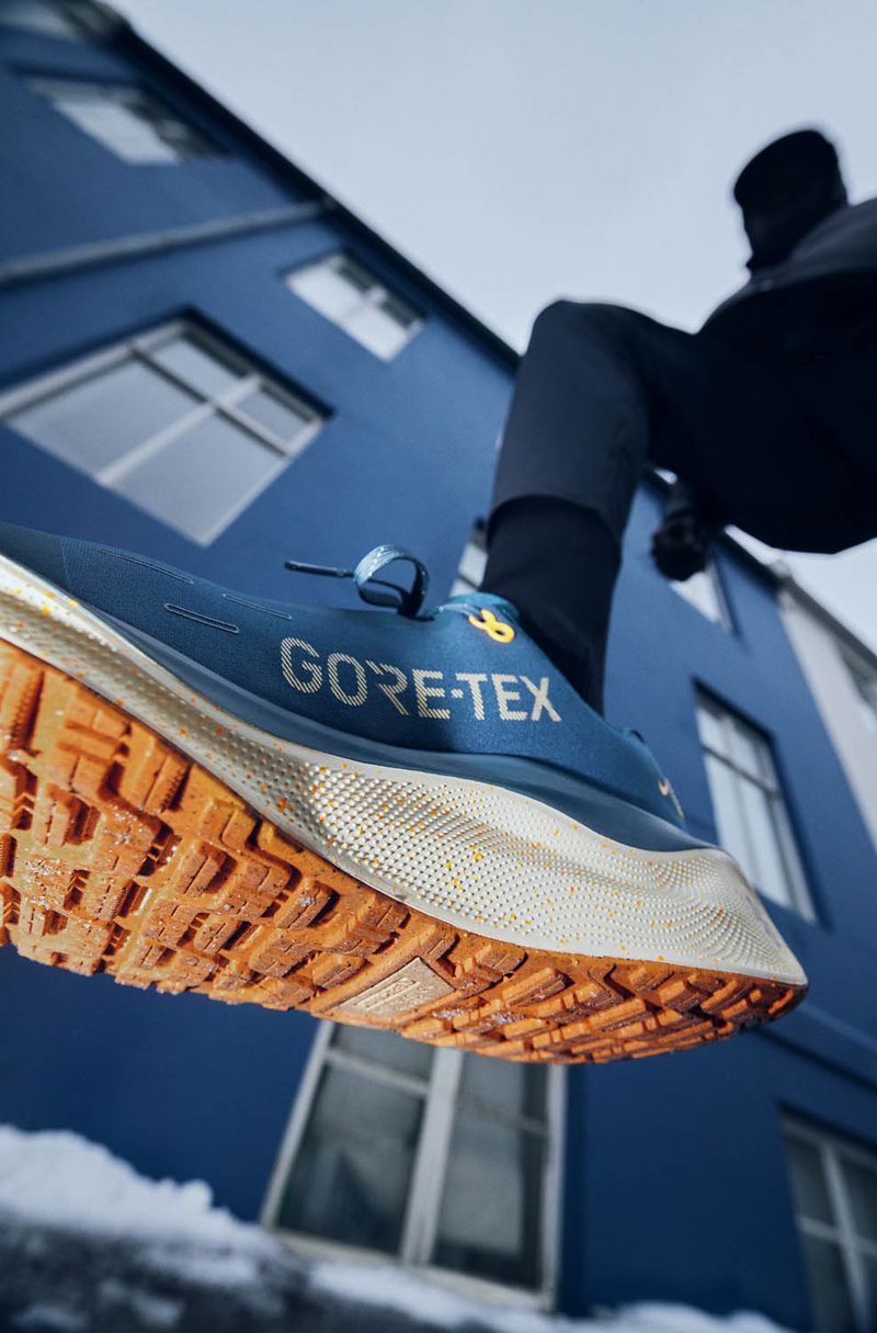 Close-up of a blue GORE-TEX running shoe with an orange sole, photographed from a low angle against a blue building and overcast sky.