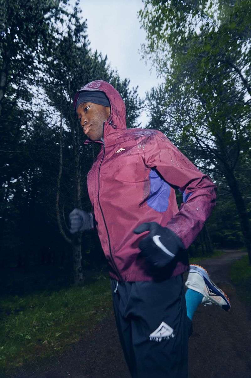 An adult male trail runner wearing a maroon hooded jacket and black athletic pants runs on a trail lined by trees.