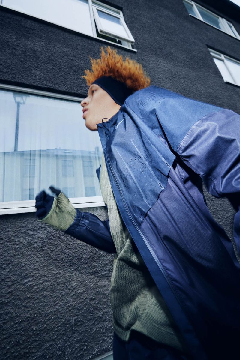 Adult runs outdoors wearing a blue and purple rain jacket, black headband, and grey gloves, captured in urban setting with cool tones.