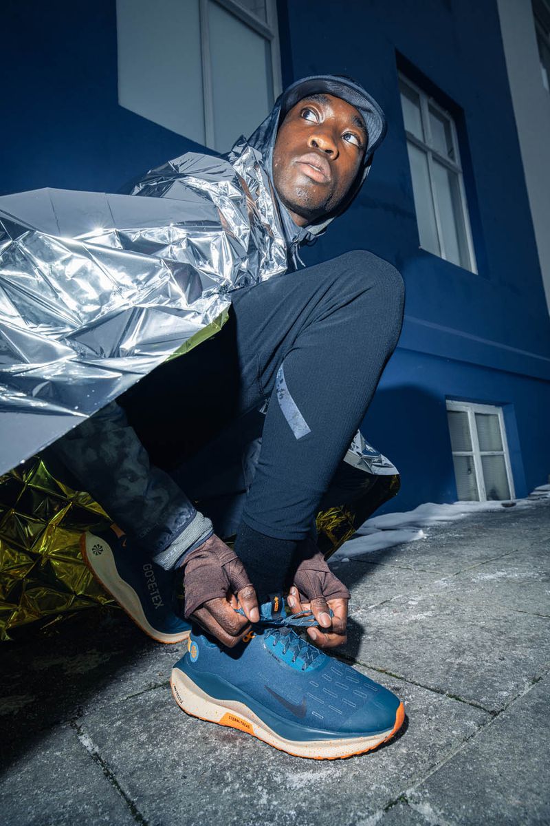 An athletic man in sportswear tying his blue running shoes outdoors at dusk. The man is wearing a reflective rain poncho.