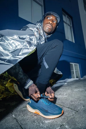 An athletic man in sportswear tying his blue running shoes outdoors at dusk. The man is wearing a reflective rain poncho.