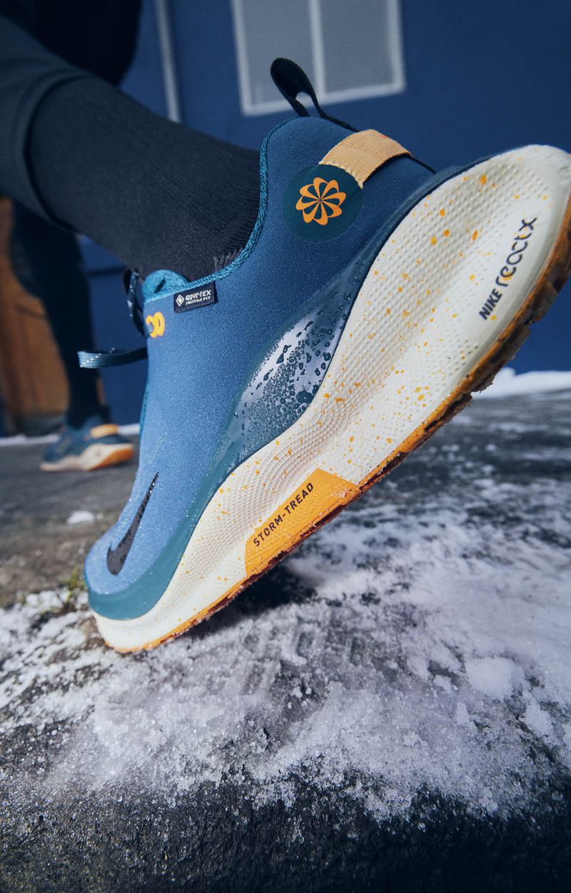 Close-up of a blue and tan Nike React GTX shoe with snowy ground. Shoe details show a Gore-Tex logo and Storm-Tread text. Footwear.