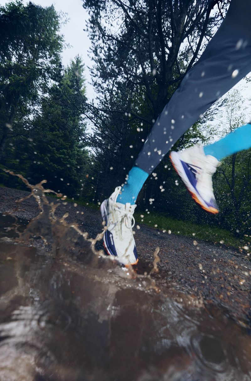 Action shot of a person running through a muddy puddle in a forest, legs and feet visible, splashing water, athletic wear, trees in background.