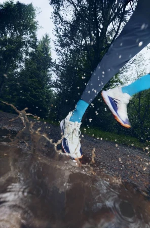 Action shot of a person running through a muddy puddle in a forest, legs and feet visible, splashing water, athletic wear, trees in background.
