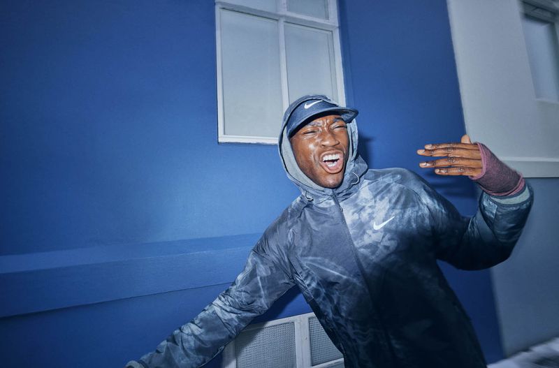 Athletic adult man in a hooded Nike jacket and hat outdoors, facial expression open, against a blue urban exterior wall with a window.