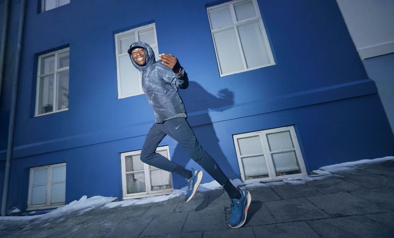 Man in athletic wear runs in an urban setting. He wears a hooded jacket and athletic pants, running on stone pavers near a blue building.