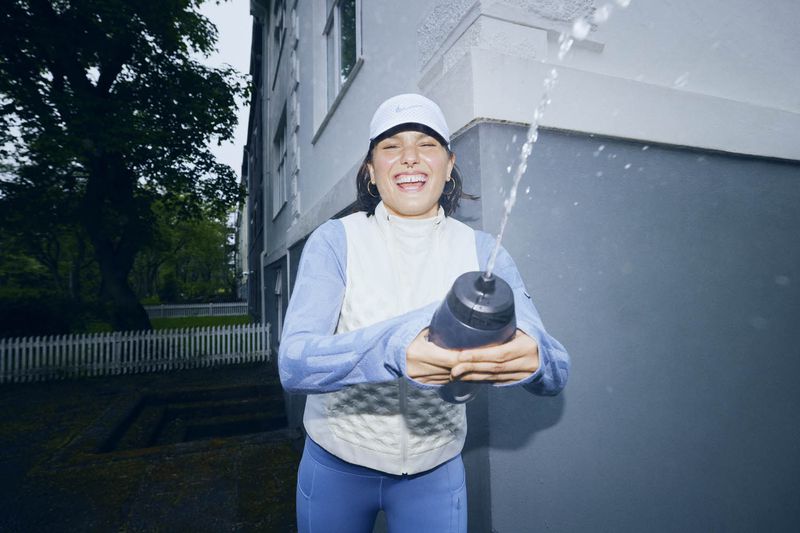Woman in athletic wear outside a building, smiling as she squirts water from a bottle, wearing a white cap, long-sleeved shirt, and leggings.