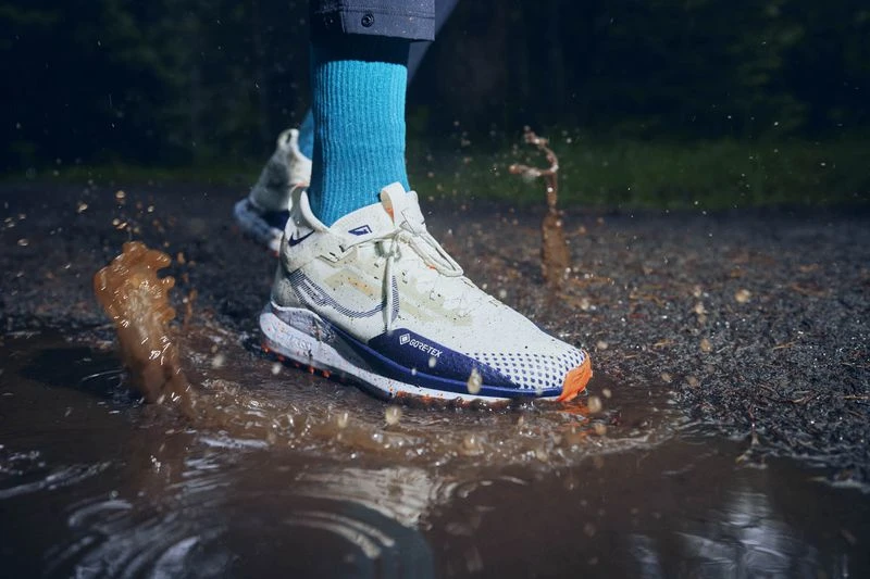 Action shot of a person's leg and Nike sneaker splashing in a muddy puddle, showcasing athletic footwear in a nature setting.