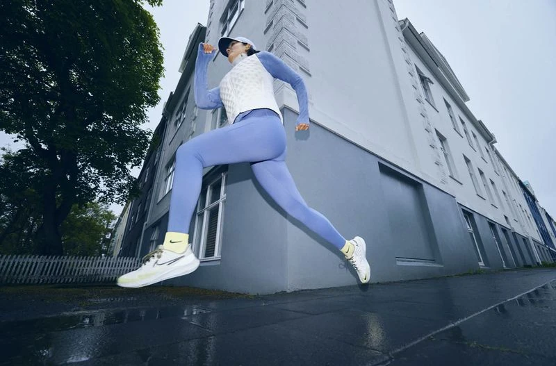 Athletic woman in a blue and white running outfit leaps along a wet urban street with buildings and trees visible in the background.