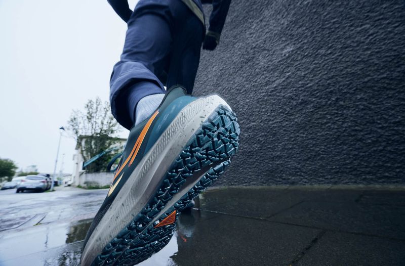 Low angle shot of a person in sportswear stepping into a puddle; showcasing the sole of a blue and orange running shoe on wet pavement.