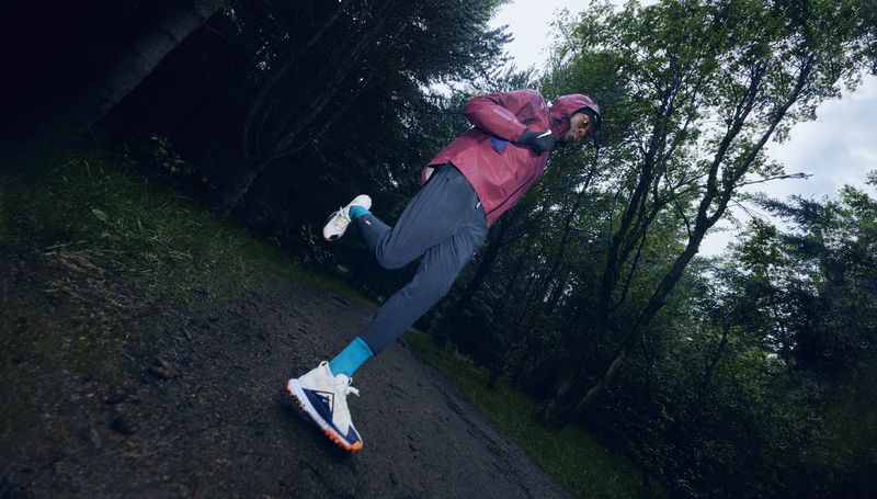 Athletic man running on a trail in a forest, wearing a burgundy jacket, blue pants, and running shoes, trees in the background.