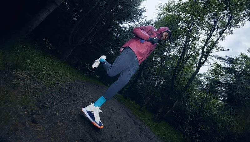 Athletic man running on a trail in a forest, wearing a burgundy jacket, blue pants, and running shoes, trees in the background.