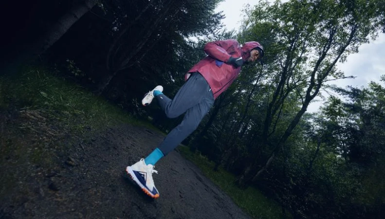 Athletic man running on a trail in a forest, wearing a burgundy jacket, blue pants, and running shoes, trees in the background.