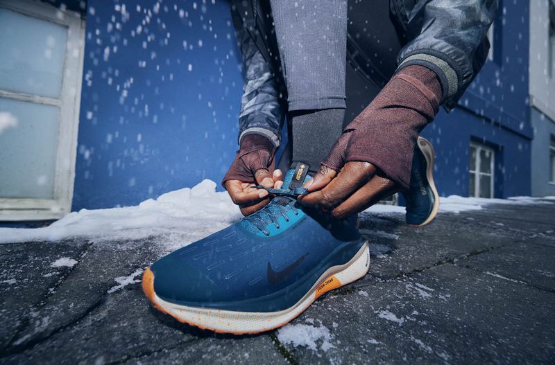 Close-up of a person tying the laces of a blue and black Nike running shoe outdoors in snowy conditions, near a blue building.