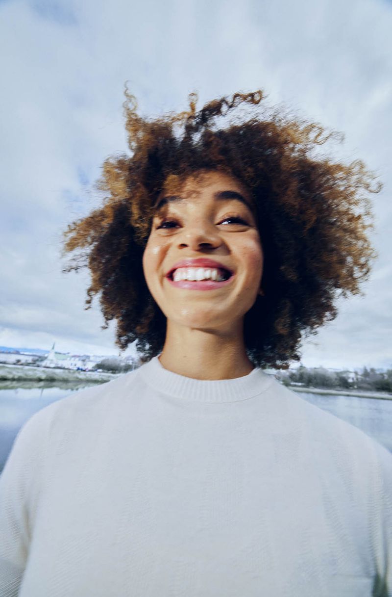 Close-up of a smiling young woman with curly hair wearing a white shirt with a landscape and cloudy sky in the background.