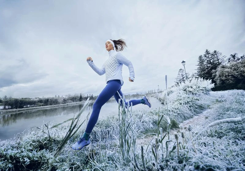 Athletic woman in sportswear running through a frost-covered landscape near a river on a cloudy winter day for fitness and wellness.