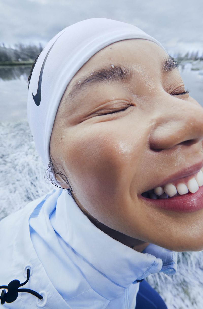 Smiling Asian woman with closed eyes wearing a Nike headband and light blue jacket near water, showcasing fitness and a positive attitude.