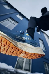 Low-angle close-up of a blue Nike Gore-Tex running shoe with an orange tread sole worn by a person mid-step in front of a blue building.