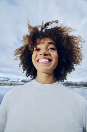 A smiling woman with curly hair looking down towards the camera wearing a white crewneck sweater outdoors near water.