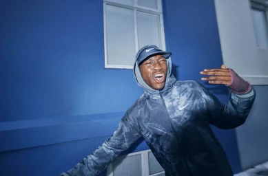 Black man wearing a hooded Nike jacket and cap making an expressive gesture in front of a blue building exterior.