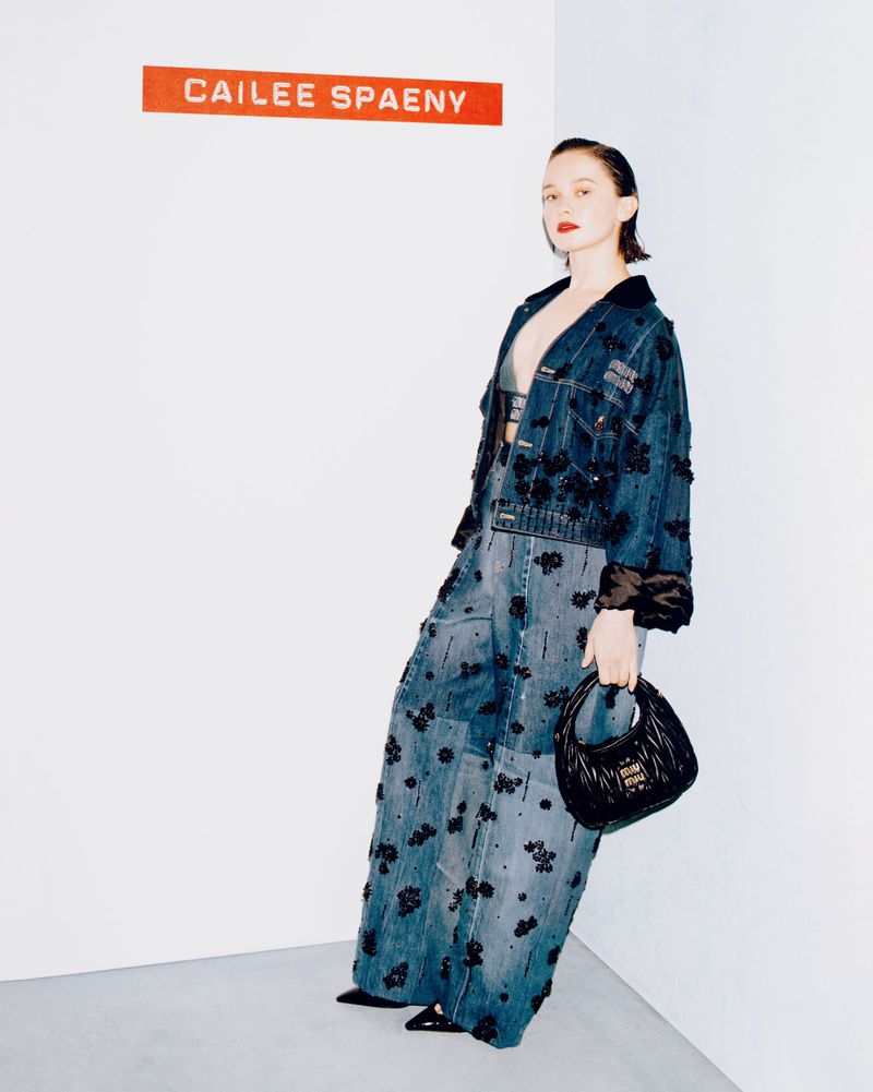 A young woman poses in a photo studio wearing a denim jacket and pants with black floral embellishments, holding a small black purse.