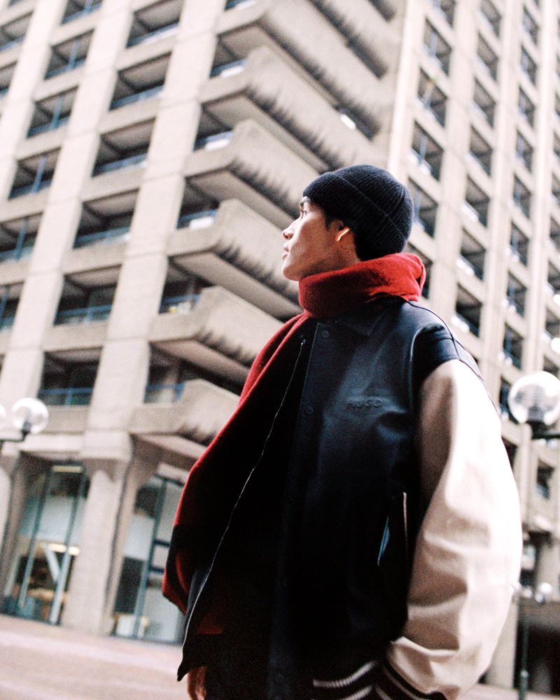 Man in a black and cream varsity jacket, vibrant red scarf, and black beanie looking up at a Brutalist residential tower.