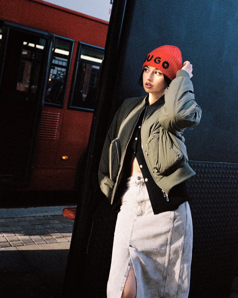 A young woman in stylish urban attire poses against a dark wall with a red bus in the background, casting a dynamic shadow.