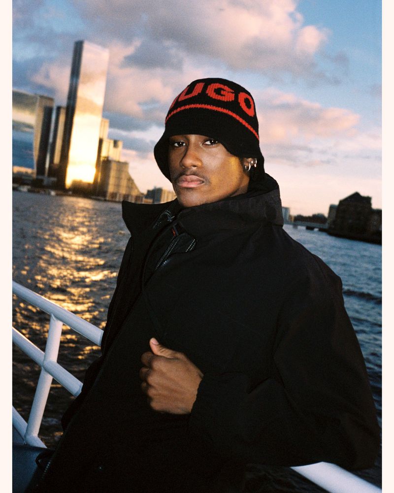 Outdoor portrait of an African-American male in a black coat and Hugo-branded hat, posing against a backdrop of city buildings and a waterway.