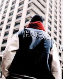 Rear view of a person wearing a black and beige HUGO leather varsity jacket, red hoodie, and black beanie in front of a tall building.