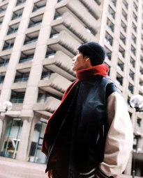 Man in a black and cream varsity jacket, vibrant red scarf, and black beanie looking up at a Brutalist residential tower.