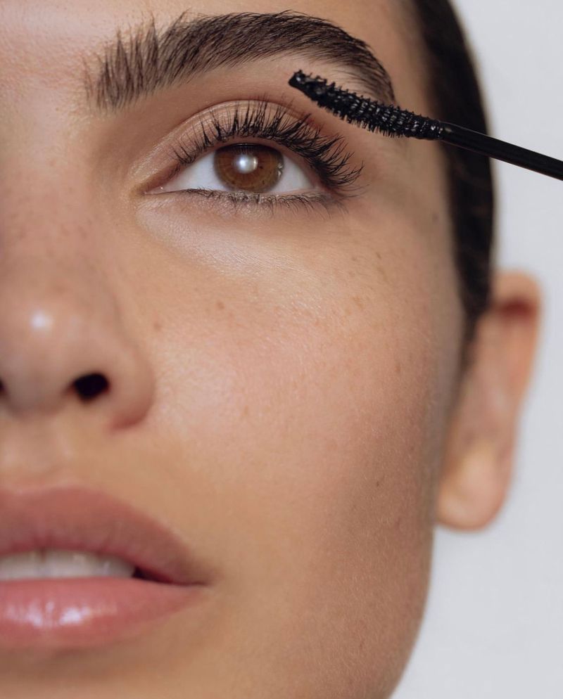 Close-up of a woman applying mascara. Beauty shot showcasing eye makeup with a wand applicator in front of a light neutral backdrop.