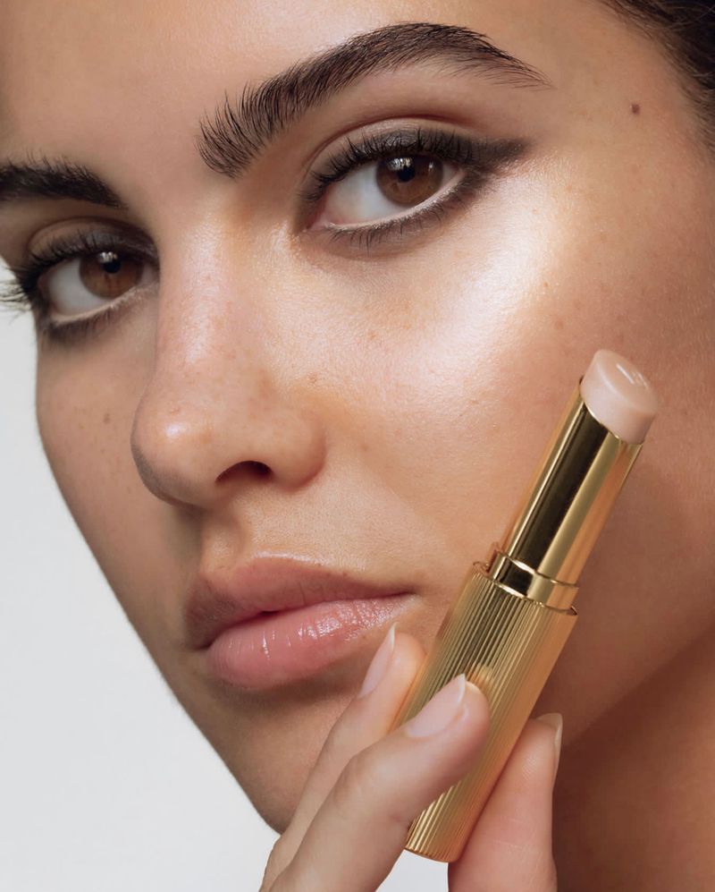 Close-up of a woman with dark hair and defined eyebrows applying lip balm, highlighting her radiant complexion and minimalist eye makeup.