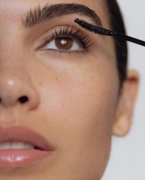 Extreme close-up of a woman with defined brows and glossy lips having black mascara applied to her upper eyelashes against a pale background.